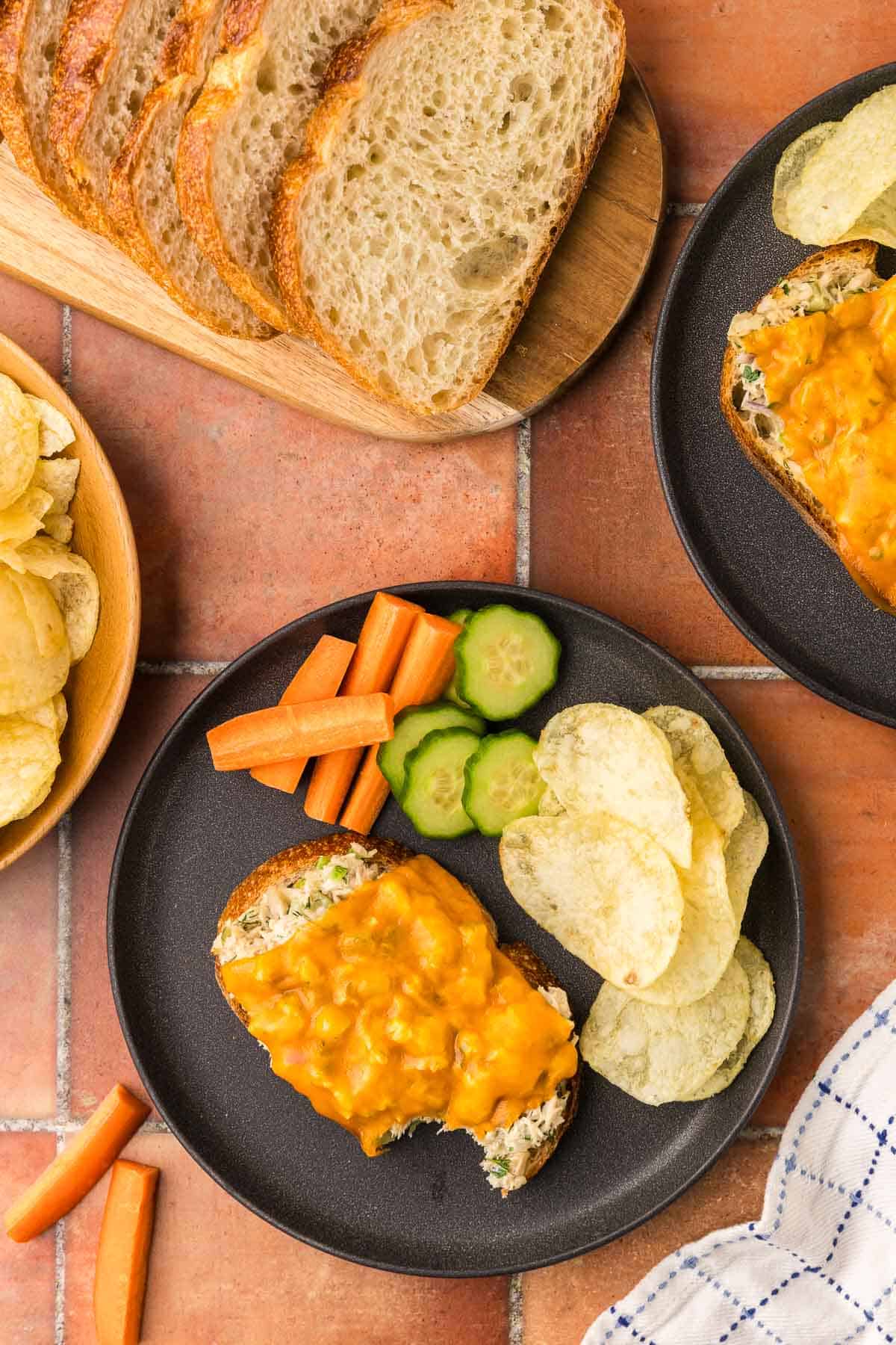 Overhead shot of a dark plate with a n open faced tun melt on it with a bite taken out of the corner. The plate also has potato chips and chopped up veggies. Another tuna melt plate is peeking in to the scene in the top right corner.