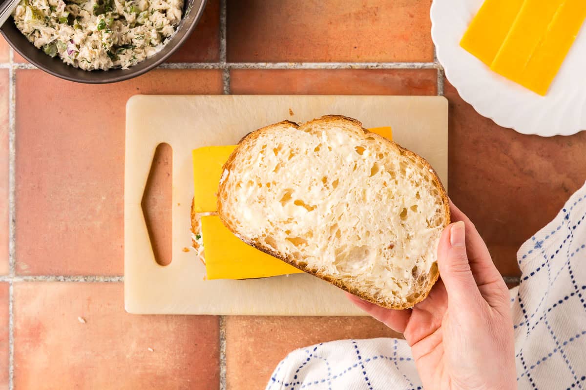 overhead shot of the final process of assembling the tuna melt. Cheese has been added and now the buttered slice of bread is placed on top to complete the sandwich.