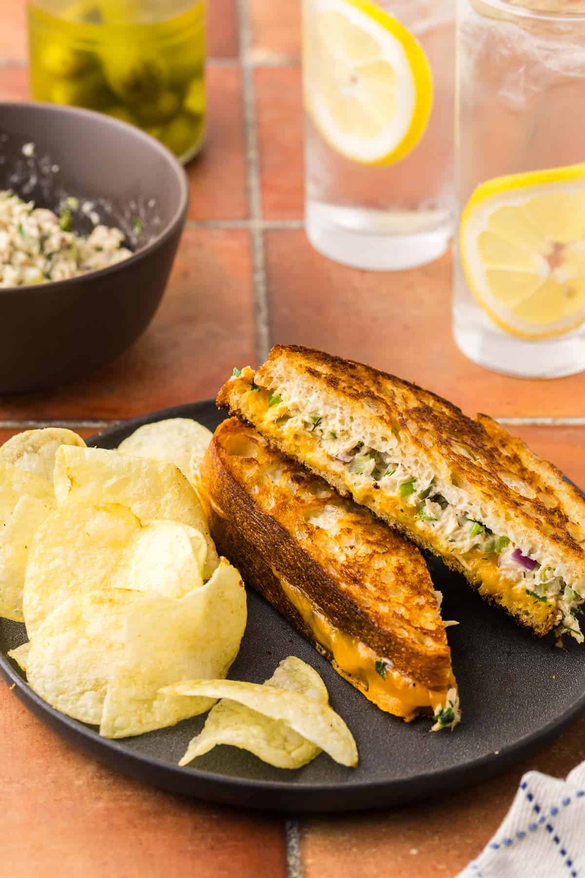 angle shot of a grilled cheese style tuna melt sliced in half on the diagonal and layed askew on a dark plate with potato chips. The cheese layer is not super cheesy for those who don't like the cheese to overpower the tuna melt.