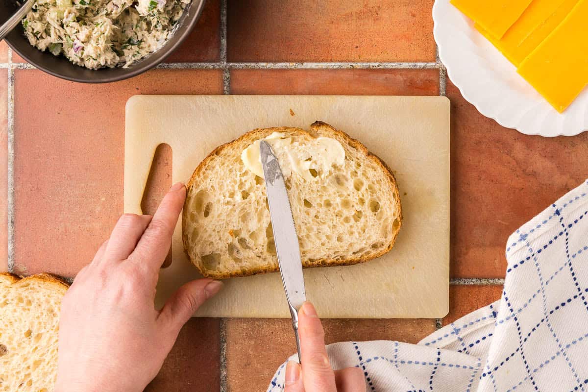 overhead shot of the initial process of making a tuna melt grilled cheese style. The bread is being buttered on the outside.