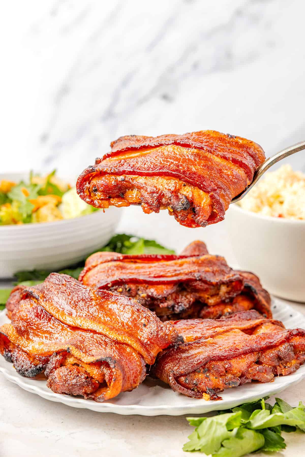 Side shot of bacon wrapped chicken thighs on a plate. One is being lifted up with a serving spoon towards the camera.