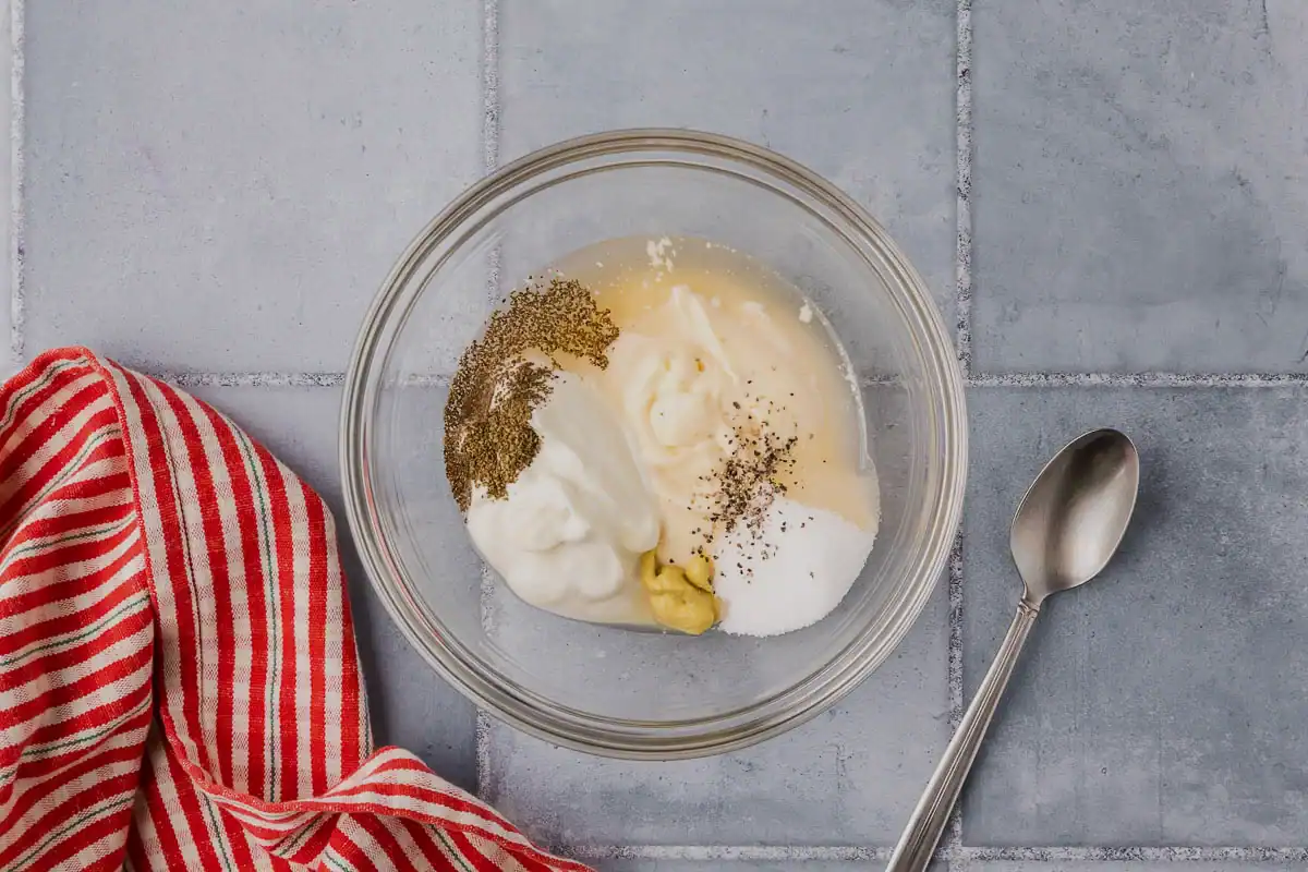 Overhead picture of a small glass bowl with all the ingredients added to make the coleslaw dressing. There is mayonnaise, Greek yogurt, apple cider vinegar, celery seed, dijon mustard, sugar and salt and pepper.