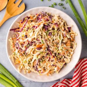 Overhead shot of a white serving bowl filled with creamy and crunchy coleslaw. Green and red cabbage make up the majority of the salad with bits of shredded carrot and a creamy dressing added. Green onions and wooden serving spoons sit in the background.