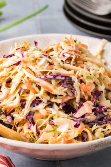 A side shot of a white serving dish filled with a creamy coleslaw. Both green and red cabbage are used and shredded carrot can be seen througout.