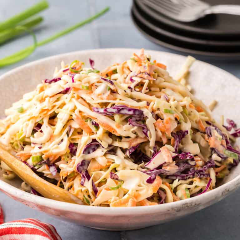 A side shot of a white serving dish filled with a creamy coleslaw. Both green and red cabbage are used and shredded carrot can be seen througout.