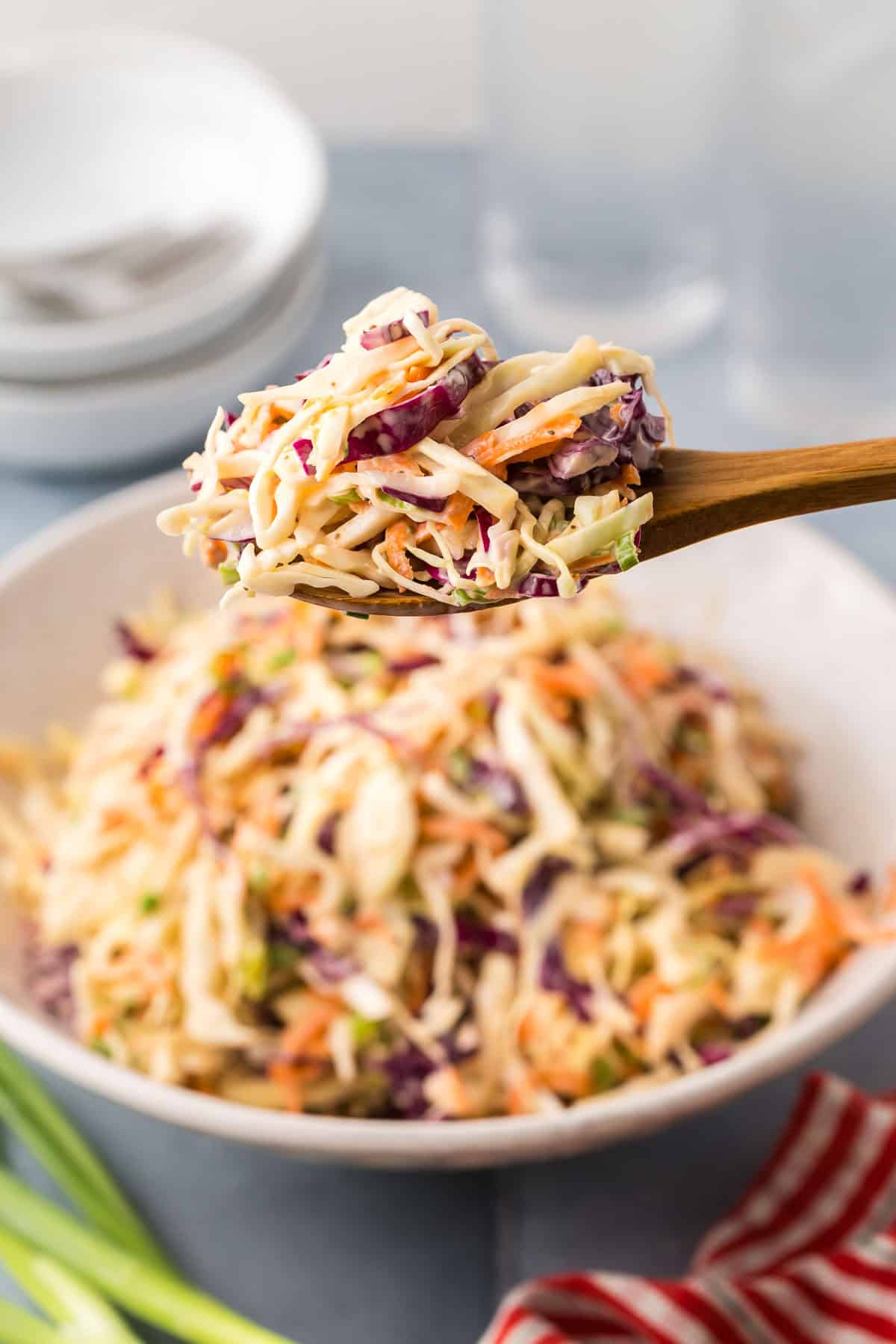 side shot focused on a close up of a serving spoon holding a scoop of shredded creamy coleslaw. Green cabbage and red cabbage are seen along with shredded carrots and sliced green onions.