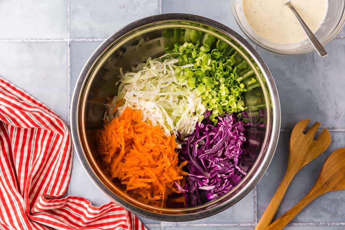 Overhead shot into a large metal mixing bowl. Inside are all the vegetables used in making coleslaw. There is green and red cabbage, shredded carrot, and sliced green onions.