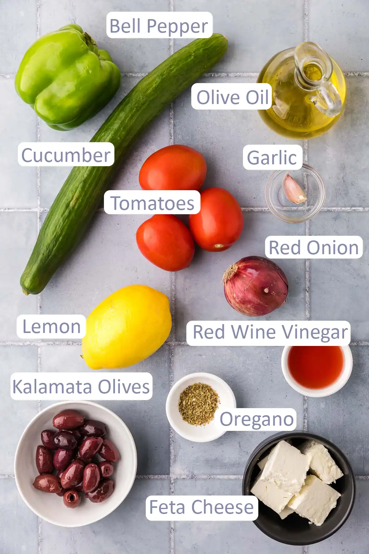 Overhead shot of all the ingredients used in making a Greek Salad. Ingredients are: bell pepper, cucumber, tomatoes, olive oil, garlic, red onion, lemon, red wine vinegar, oregano, kalamata olives, and feta cheese.