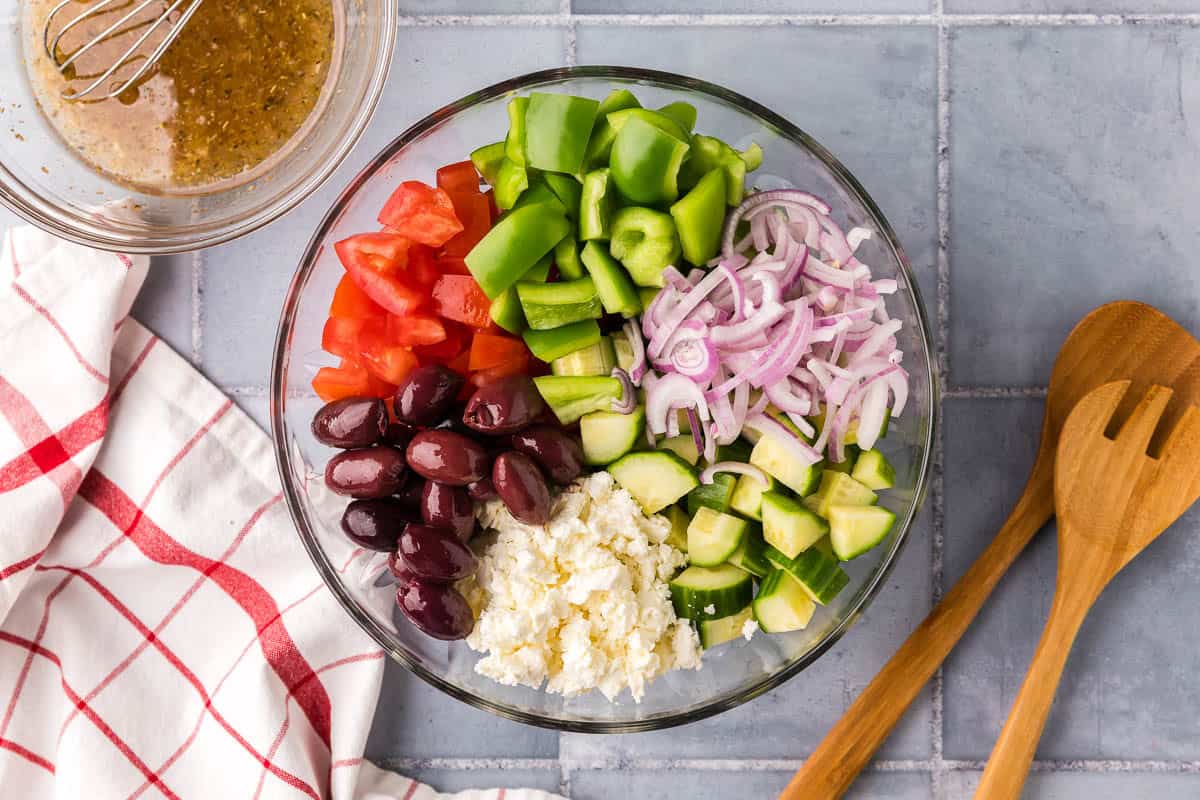 Overhead shot of a glass mixing bowl filled with piles of chopped green peppers, sliced red onions, chopped cucumber, crumbled feta cheese, kalamata olives, and chopped tomatoes.