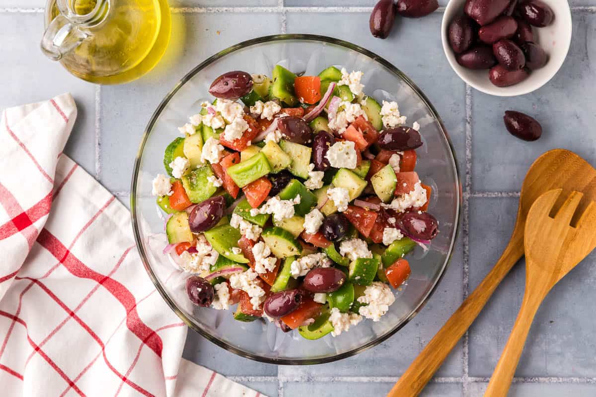 overhead shot of a glass mixing bowl filled with Greek Salad. Lots of tomatoes, cucumbers, green peppers, kalamata olives and feta can be seen.