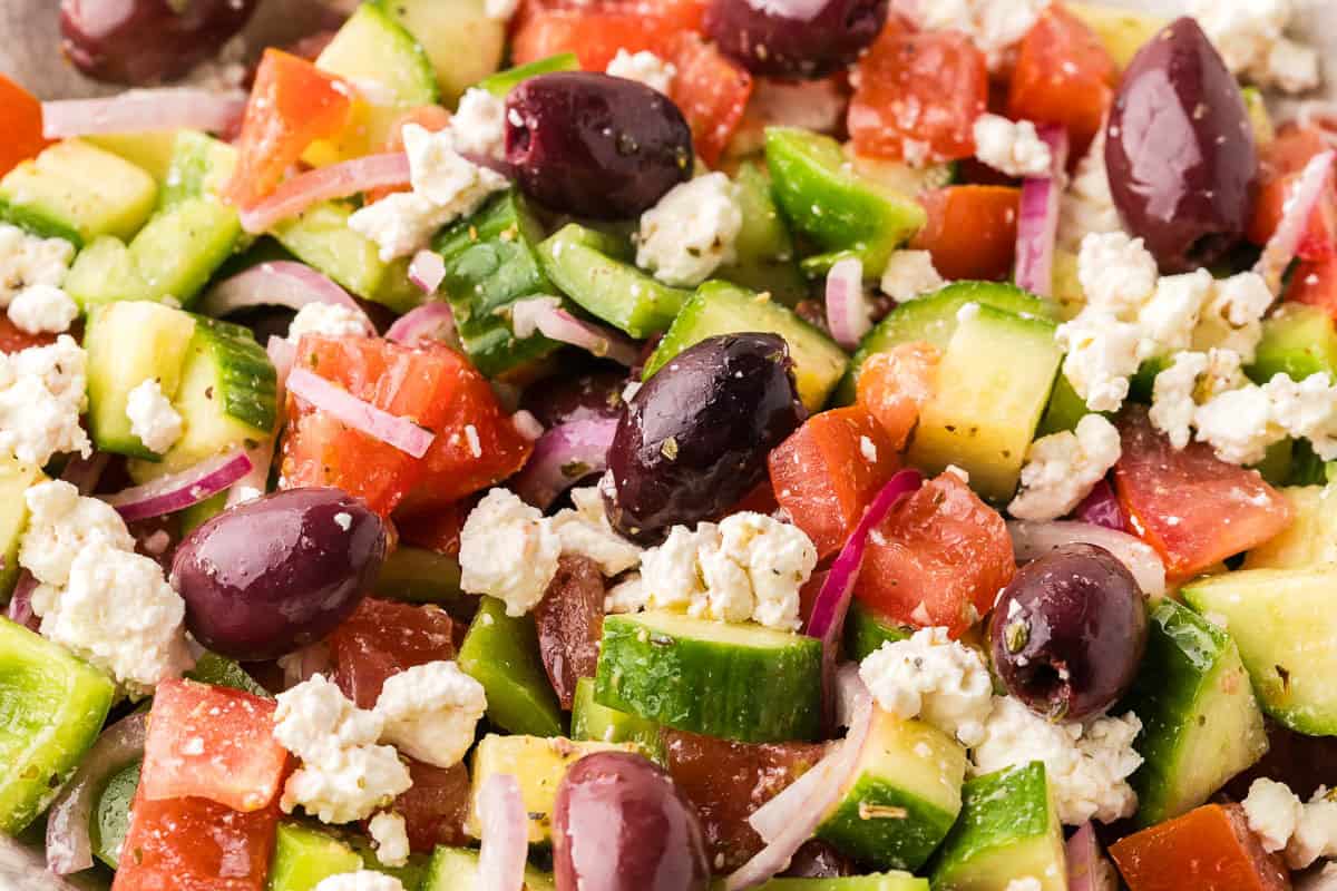 Close up shot looking into a bowl of Greek salad. The whole screen is taken up by chunks of tomatoes, green bell peppers, sliced onions, kalamata olive, feta cheese, all in a shiny dressing.