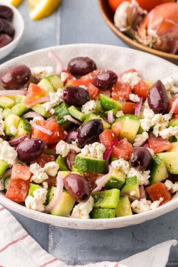 side shot of a white serving bowl filled with Greek Salad. Lots of tomatoes, cucumbers, green peppers, kalamata olives and feta can be seen. In the background is a wooden bowl of fresh tomatoes and onions and a small bowl of dark kalamata olives.