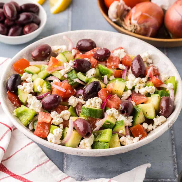 side shot of a white serving bowl filled with Greek Salad. Lots of tomatoes, cucumbers, green peppers, kalamata olives and feta can be seen. In the background is a wooden bowl of fresh tomatoes and onions and a small bowl of dark kalamata olives.