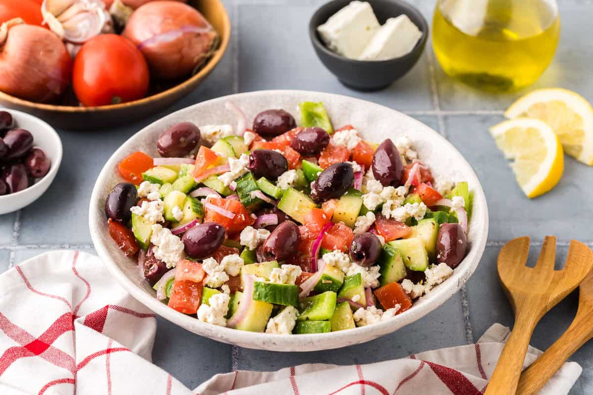 side shot of a white serving bowl filled with Greek Salad. Lots of tomatoes, cucumbers, green peppers, kalamata olives and feta can be seen. In the background is a bowl of fresh tomatoes and onions and a small bowl of feta.