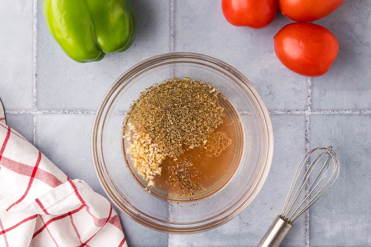 overhead shot of a small glass bowl with all the Greek salad dressing ingredients added: olive oil, red wine vinegar, lemon juice, and oregano. A whisk is laying beside the bowl.