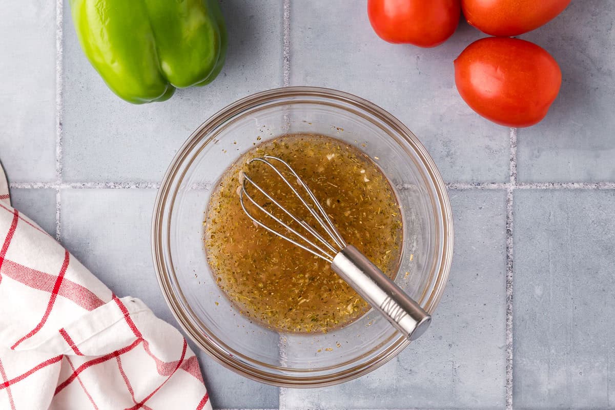 overhead shot of a small glass bowl with all the ingredients for Greek salad added and whisked together. The form a fairly homogenous brown/yellow mixture with flecks of oregano througout.