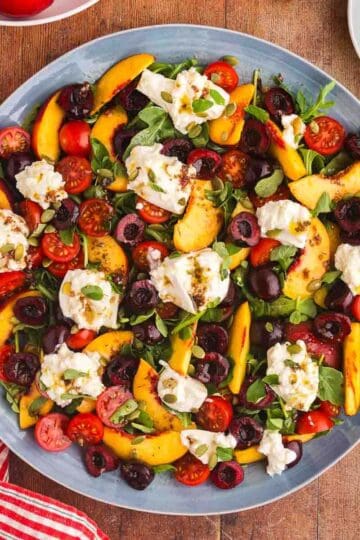 Overhead shot of a large circular blue serving plate topped with sliced fresh peaches, creamy chunks of burrata, sliced cherries, tomatoes, and fresh herbs.