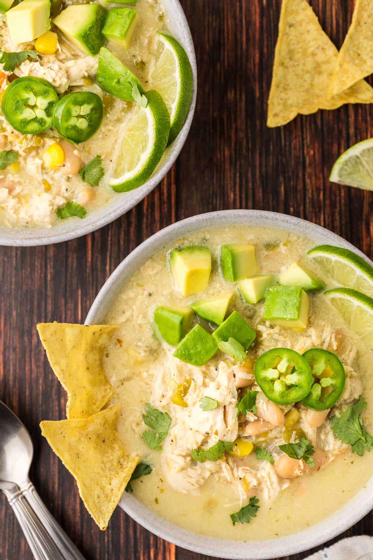 Overhead shot of a bowl of white chili chicken garnished with jalapeno slices, cilantro, chunks of avocado and two tortilla chips. Another bowl is poking in the top left corner.