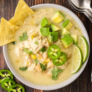 Overhead close up shot of a bowl of white chili chicken garnished with green jalapeno slices, cilantro, lime wedges, cubed avocado, and tortilla chips.