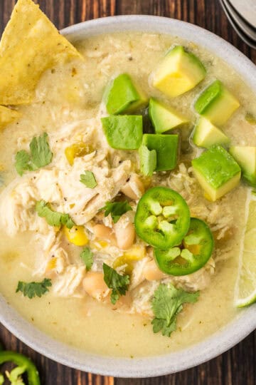 Overhead close up shot of a bowl of white chili chicken garnished with green jalapeno slices, cilantro, lime wedges, cubed avocado, and tortilla chips.