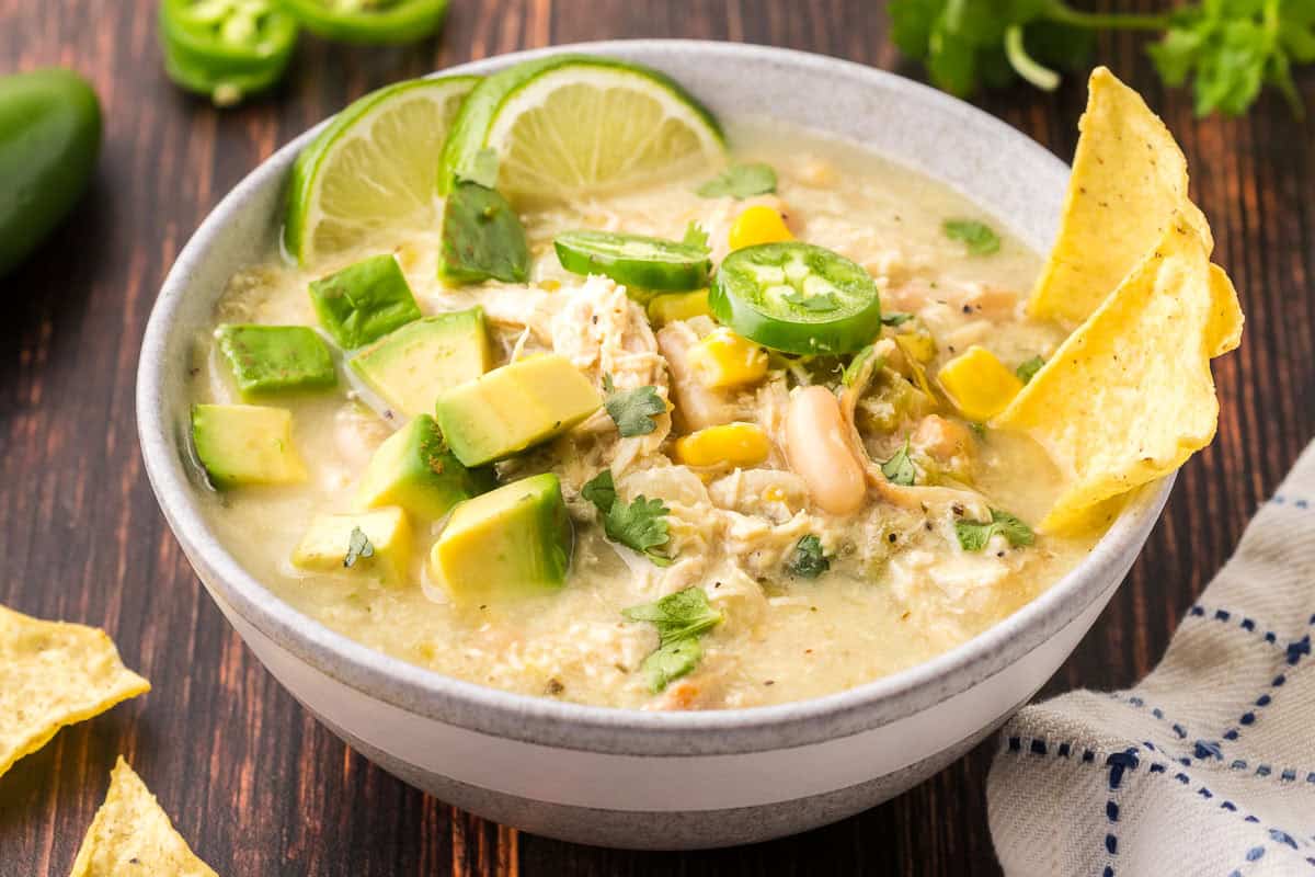 Side shot of a bowl of white chili chicken served and garnished with small chunks of avocado, lime wedges, jalapeno slices, cilantro, and a couple of tortilla chips.